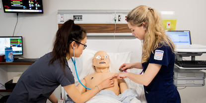 Two nursing students practicing skills in hospital over a bed with a medical simulation mannequin. 